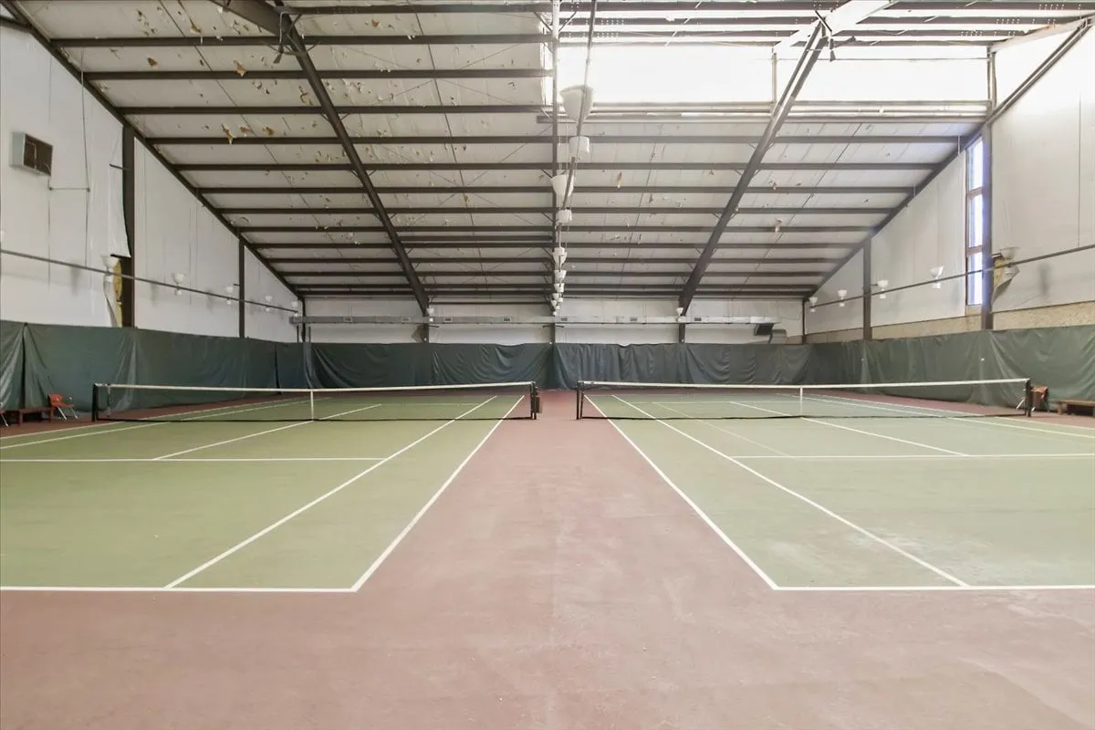 A spacious, well-lit interior tennis court with clear netting and court markings. The scene suggests energy and community activity, ideal for group matches or training sessions regardless of weather.