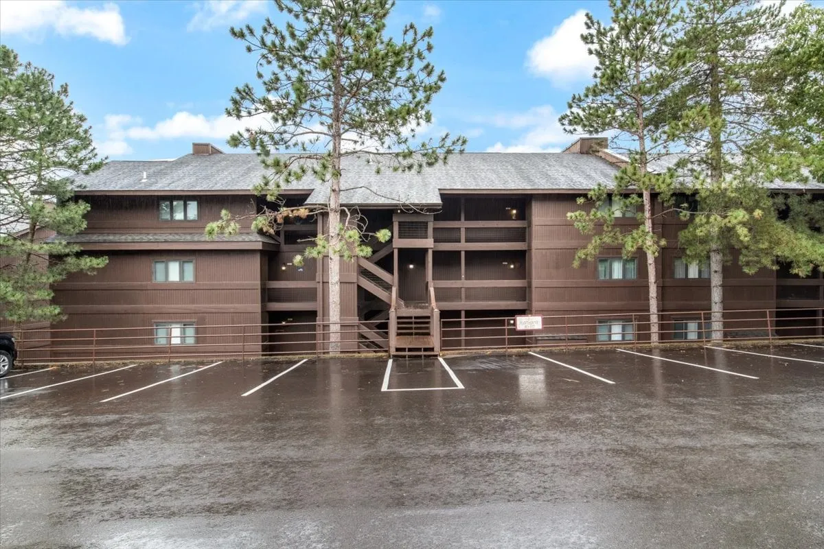 The wooden exterior of the condo building, bordered by mature trees and a spacious parking area. The setting feels peaceful and open, inviting visitors to imagine arriving for a vacation getaway.