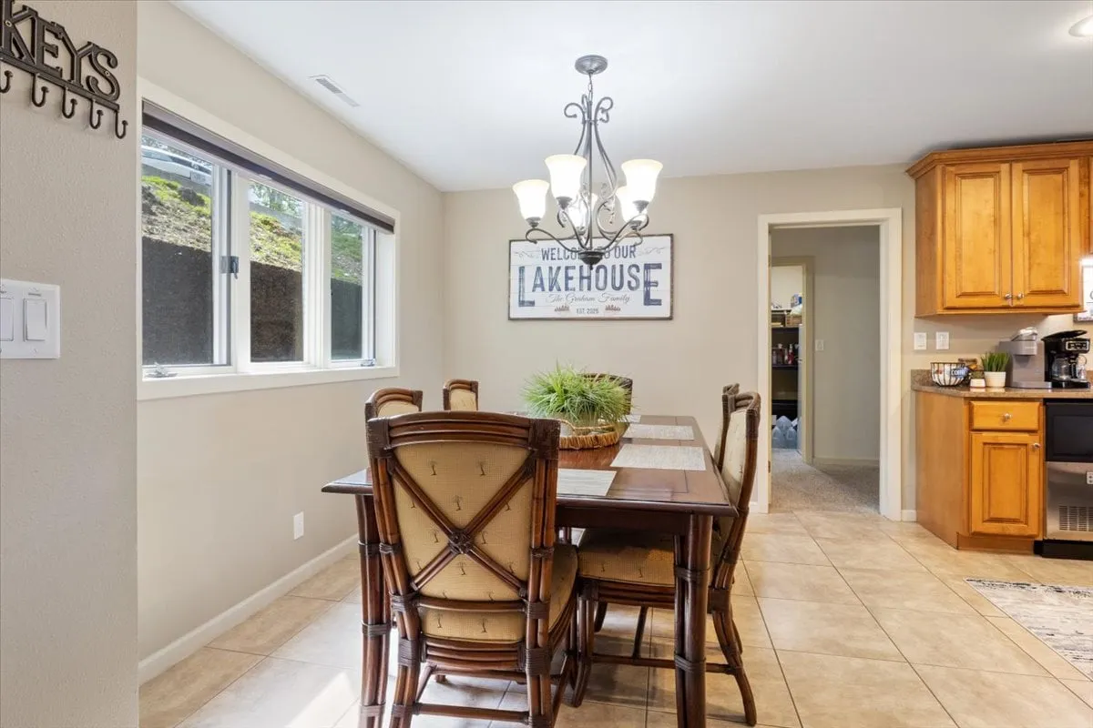 A spacious dining area with a large table, elegant chandelier, kitchen cabinetry, and a cheerful 'lakehouse' sign. The mood is festive and communal, evoking family feasts, celebratory toasts, and entertaining guests.