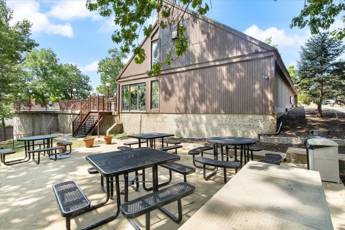 The image showcases picnic tables set under the trees beside the building’s exterior, suggesting a casual, social atmosphere. The open outdoor space invites guests to imagine group gatherings after a day on the lake or exploring the area.