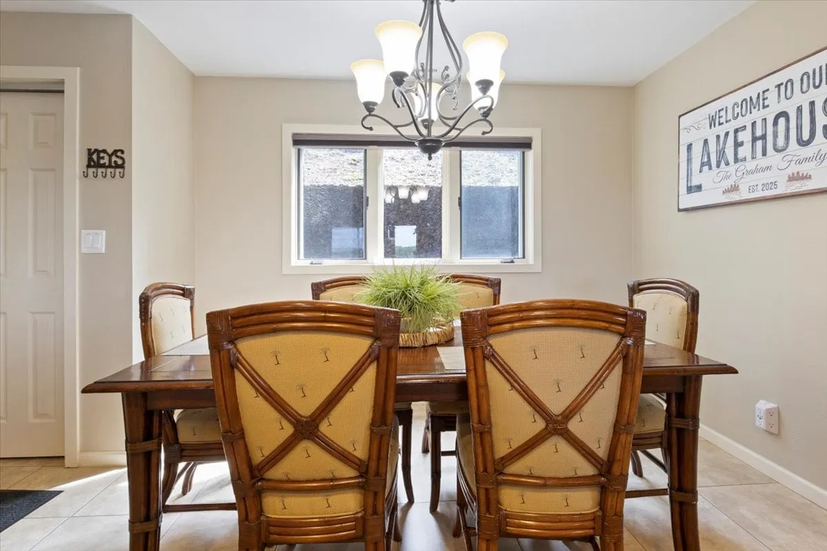 A bright, welcoming dining area featuring a spacious table under a chandelier, with a 'lakehouse' sign on the wall and large windows offering views outside. The setting is perfect for family meals, celebrations, and shared stories, with the ambiance hinting at the lakeside location.