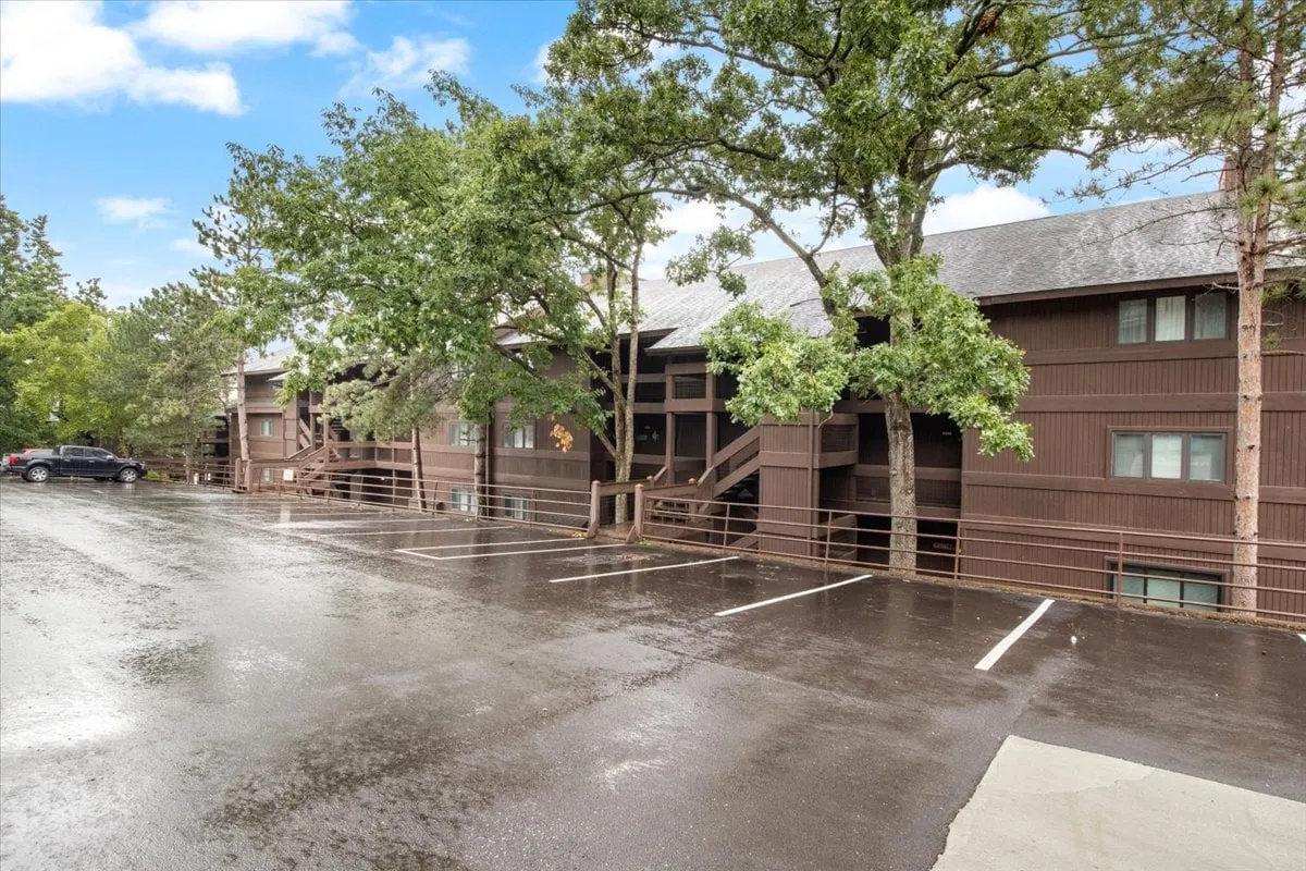 A wooden building facade surrounded by lush green trees and set against a slightly wet pavement, suggesting recent rain. The tranquil outdoor environment offers a welcoming sense of community and nature, capturing the entryway to the resort-style property.