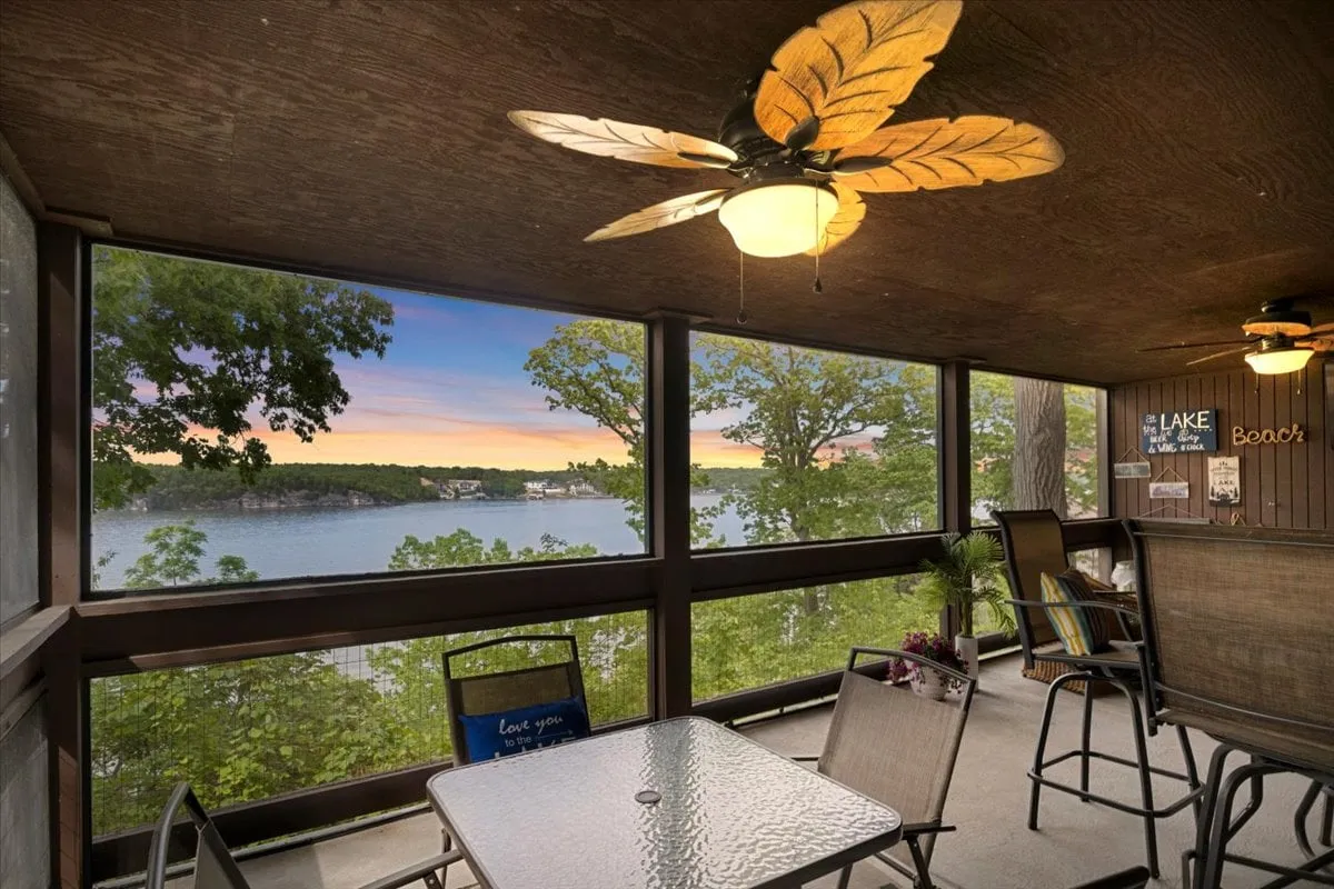 The indoor dining area is depicted with a cozy table, ceiling fan, and expansive lake views visible through the windows. The atmosphere is intimate yet open, ideal for shared meals, laughter, and memorable conversations against a scenic backdrop.