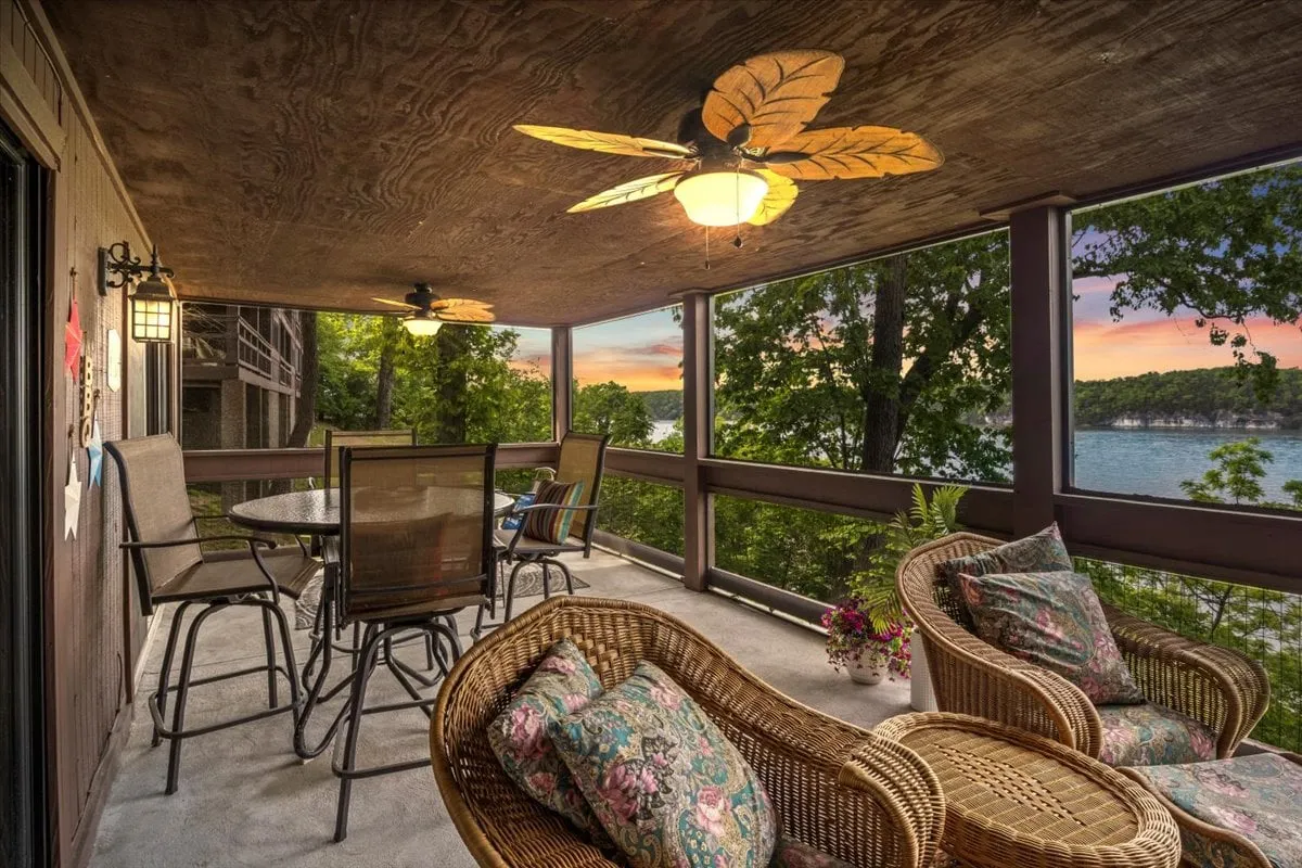 The image showcases an inviting outdoor seating area on a screened patio overlooking the tranquil lake, with a decorative ceiling fan above. The scene conveys a sense of comfort and relaxation, with the promise of stunning sunset views and cozy mornings with coffee. The lake is clearly visible, enhancing the connection to nature and offering a glimpse of the main channel.