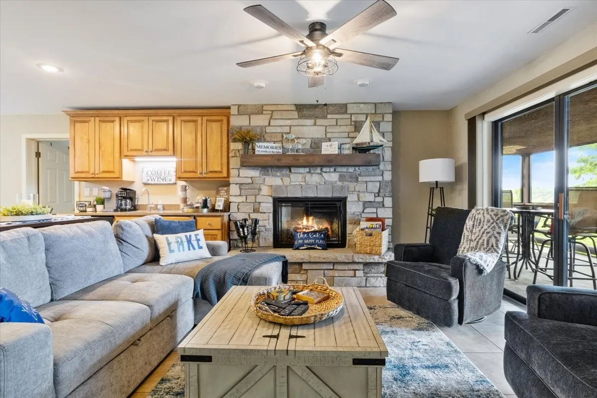 An open living area seamlessly connected to the kitchen, with a cozy fireplace, a large sofa, coffee table, and expansive windows. The layout encourages socializing, relaxing, and game nights—ideal for both quiet moments and lively get-togethers.