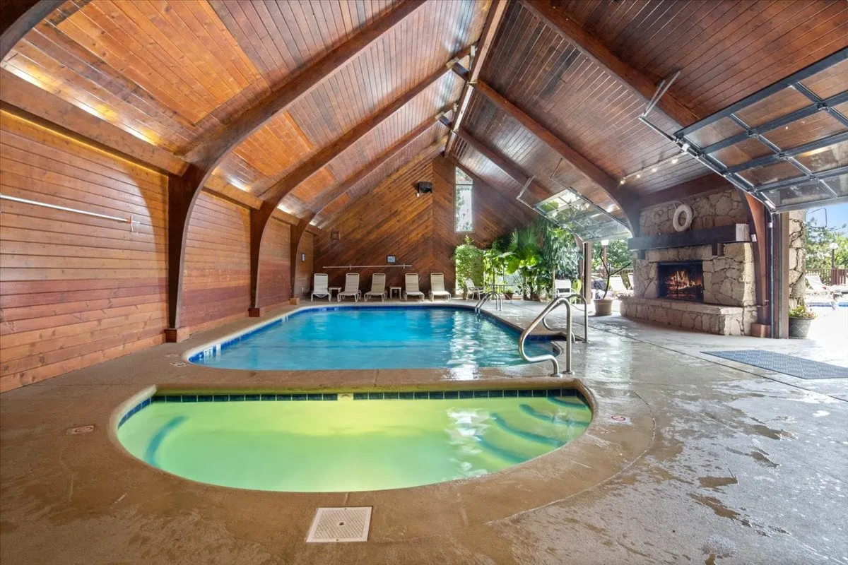 A spacious indoor pool area with a wooden ceiling, a cozy fireplace, and adjacent spa hot tub. The tranquil pool is surrounded by comfortable seating and sunbathing spots, creating a relaxing environment for swimming, soaking, and socializing year-round.