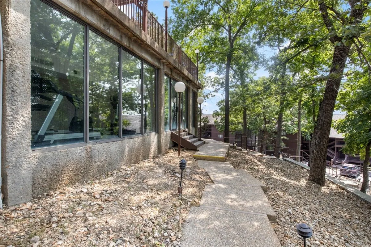 The image captures a building with large glass windows, a winding pathway, and mature trees, all bathed in natural light. The outdoor lighting and connection to nature invite guests to imagine leisurely strolls and relaxation after a day of adventure.