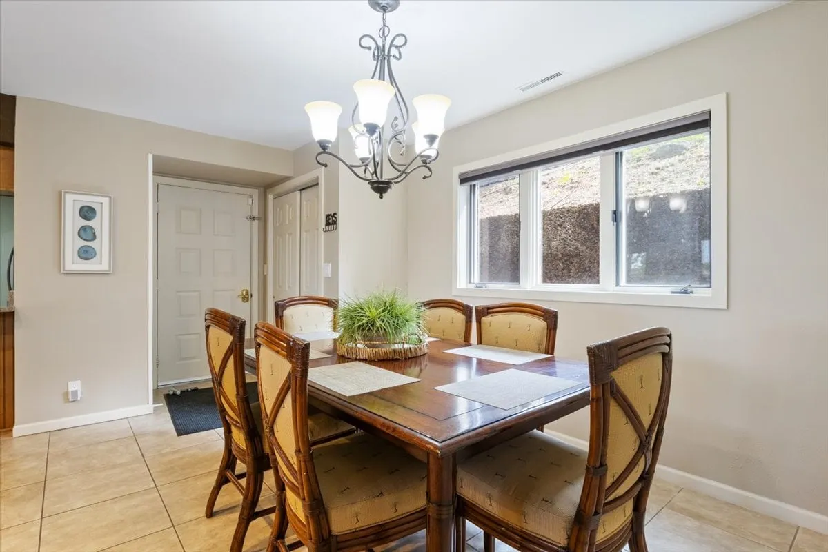 A warmly lit dining area centered around a large table with a stylish chandelier overhead and a window that lets in ample natural light. The space exudes an inviting atmosphere ideal for group meals or celebrations, with enough seating for several guests to gather comfortably.